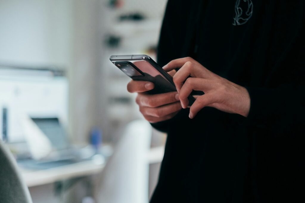 Hände halten ein Smartphone vor einem unscharfen Arbeitsplatz im Hintergrund