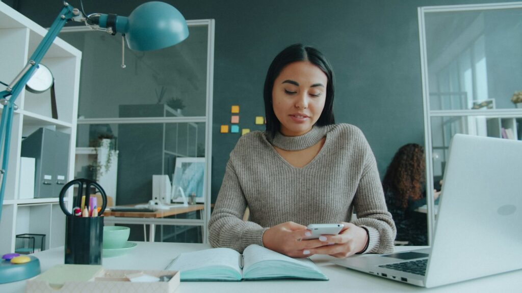 Frau prüft am Büroschreibtisch etwas auf dem Smartphone, daneben Notizbuch und Laptop
