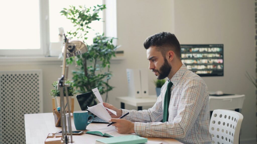 Mann prüft am Schreibtisch einen ausgedruckten Text mit Stift, daneben Laptop