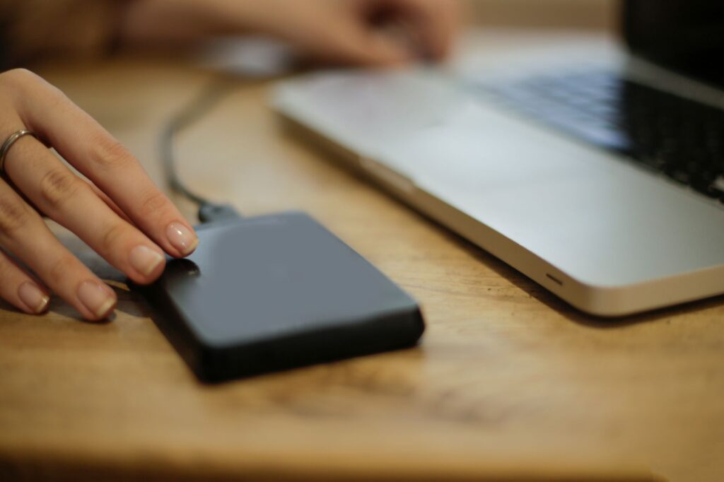 Hand an einer externen Festplatte, die per Kabel neben einem Laptop liegt, als zusätzliche Sicherung für heruntergeladene iCloud-Fotos