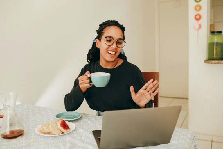 Teams Status immer verfügbar – Frau mit Kaffeetasse winkt lächelnd vor ihrem Laptop